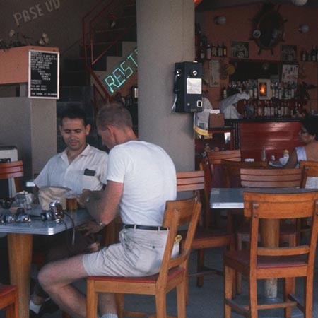 During a break from the Swan Song Expedition (1961) a member of the crew took this photo of two other crew members enjoying a waterfront café in Acapulco, Mexico. 1961.